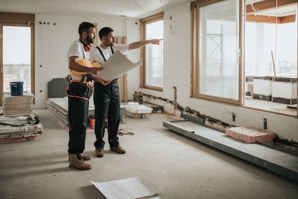 Full length of construction workers analyzing blueprints in the apartment while one of them is aiming at distance.